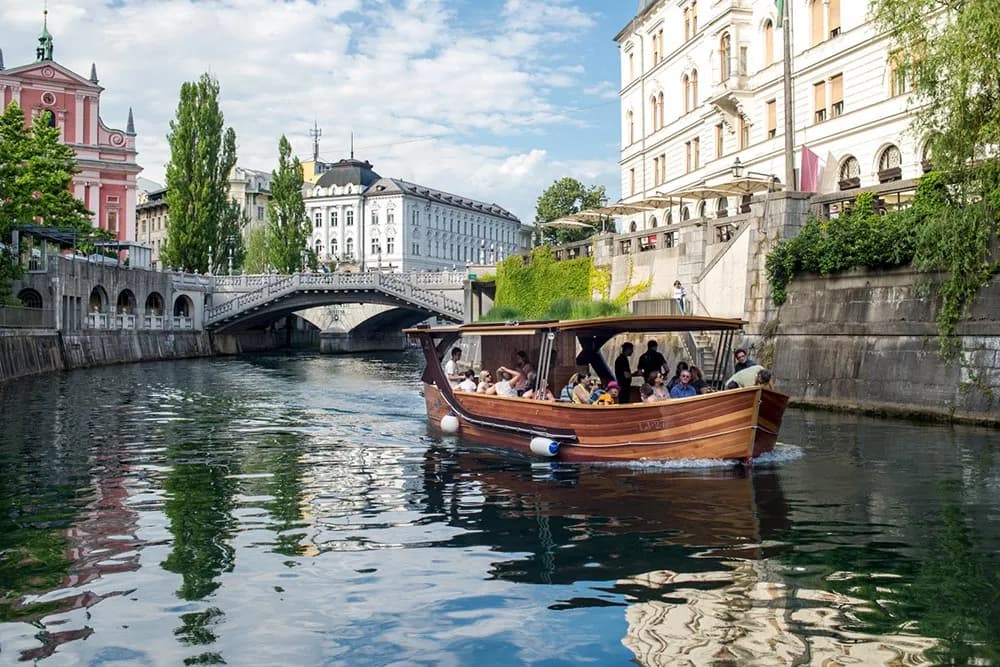 Wooden tour boat with passengers cruising the Ljubljanica River in Ljubljana past historic buildings.