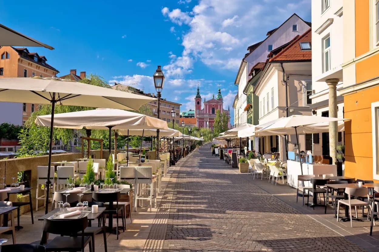 Outdoor cafe seating on cobblestone street with pink Franciscan Church in Ljubljana