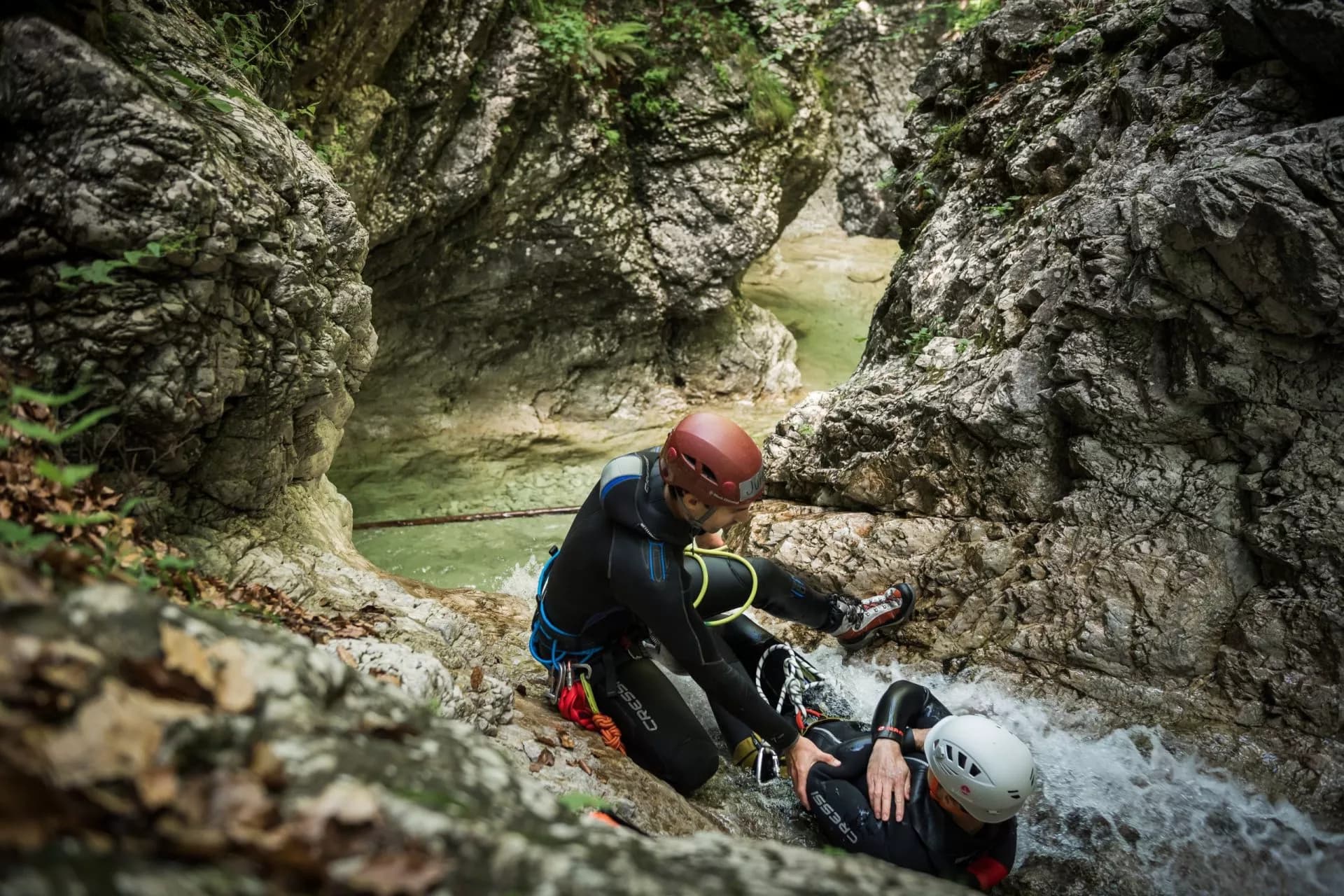 Canyoning in Slovenia: two people in wetsuits navigating a narrow, rocky gorge with flowing green water.