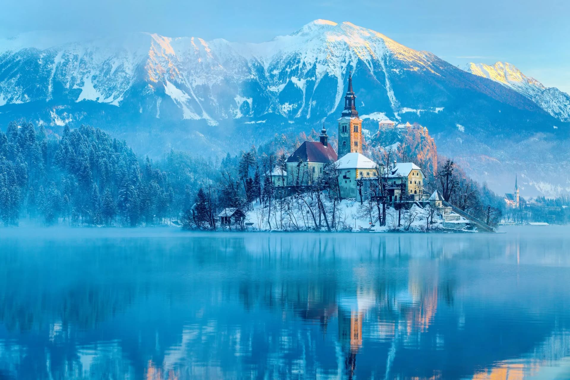 Winter scene at Lake Bled with snow-covered island church reflecting in misty blue water and mountains.