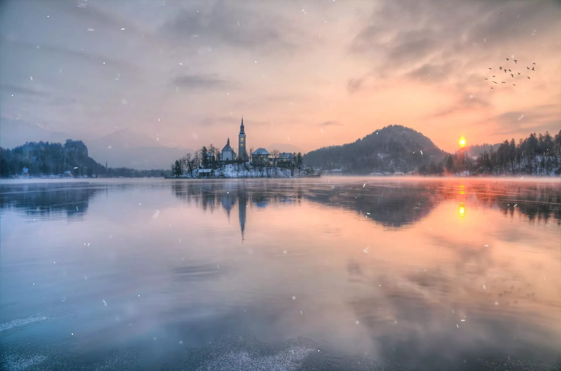 Winter sunrise at Lake Bled with island church, snow, and mist over water.
