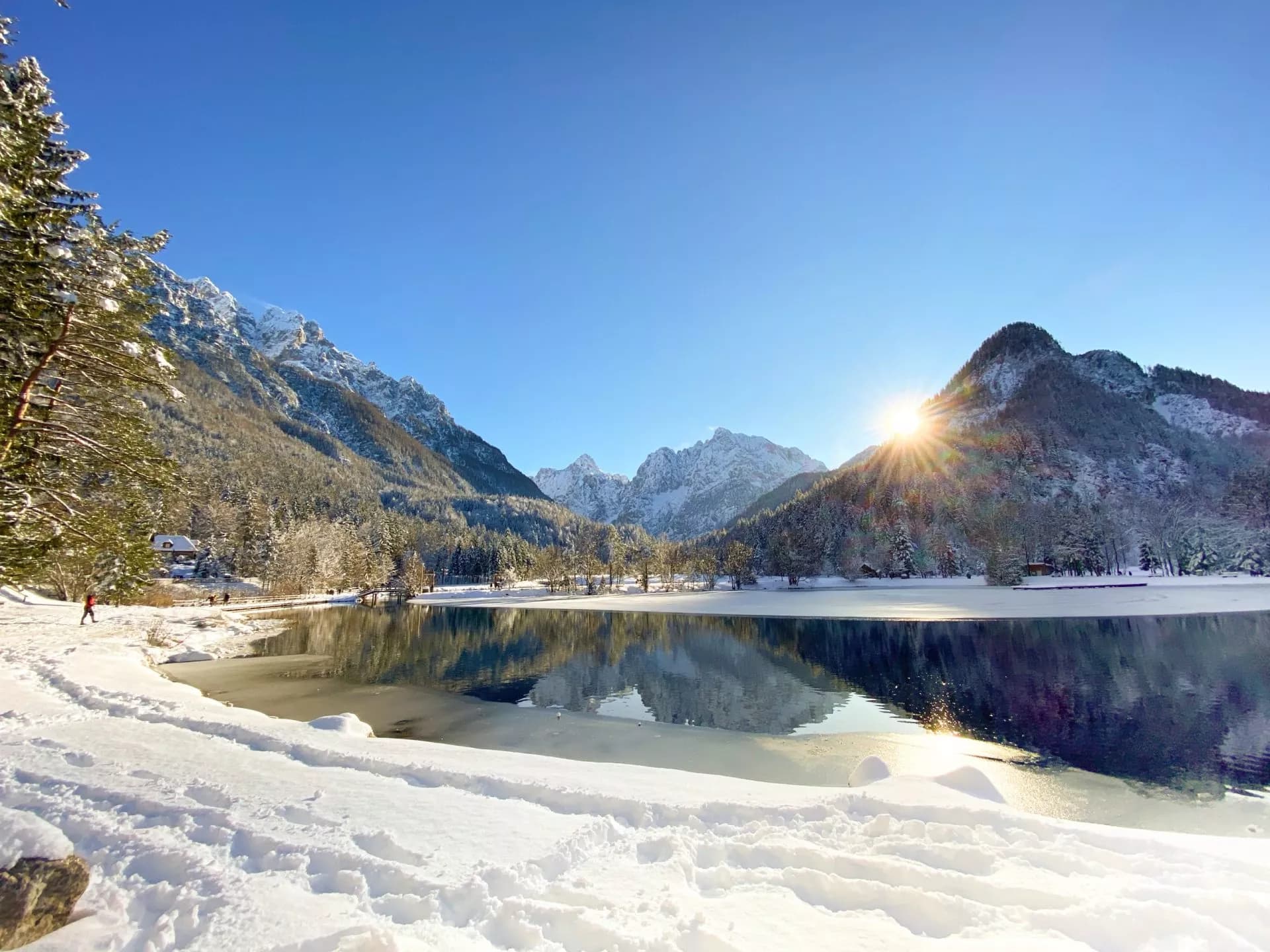 Winter sun over Lake Kranjska Gora reflecting in the partially frozen lake with snow-covered mountains.