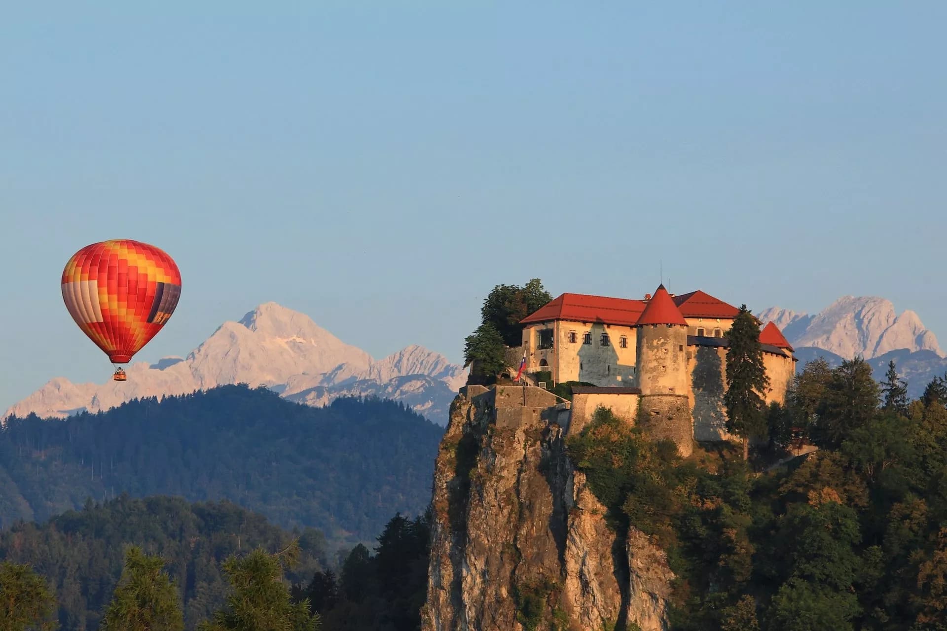 Hot air balloon flying near Bled Castle with snow-capped mountains in the background.