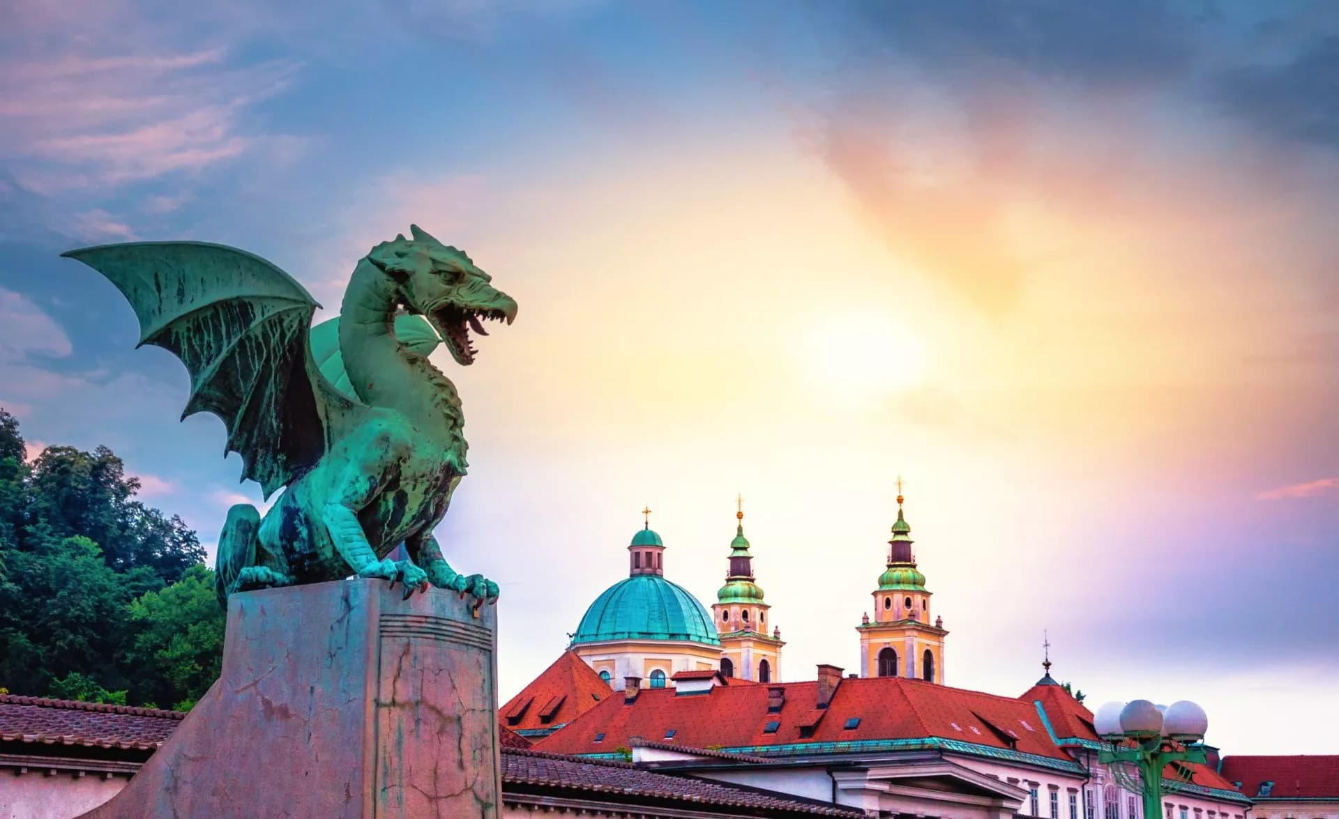 Dragon statue on Ljubljana Dragon Bridge with church domes and sunset sky.