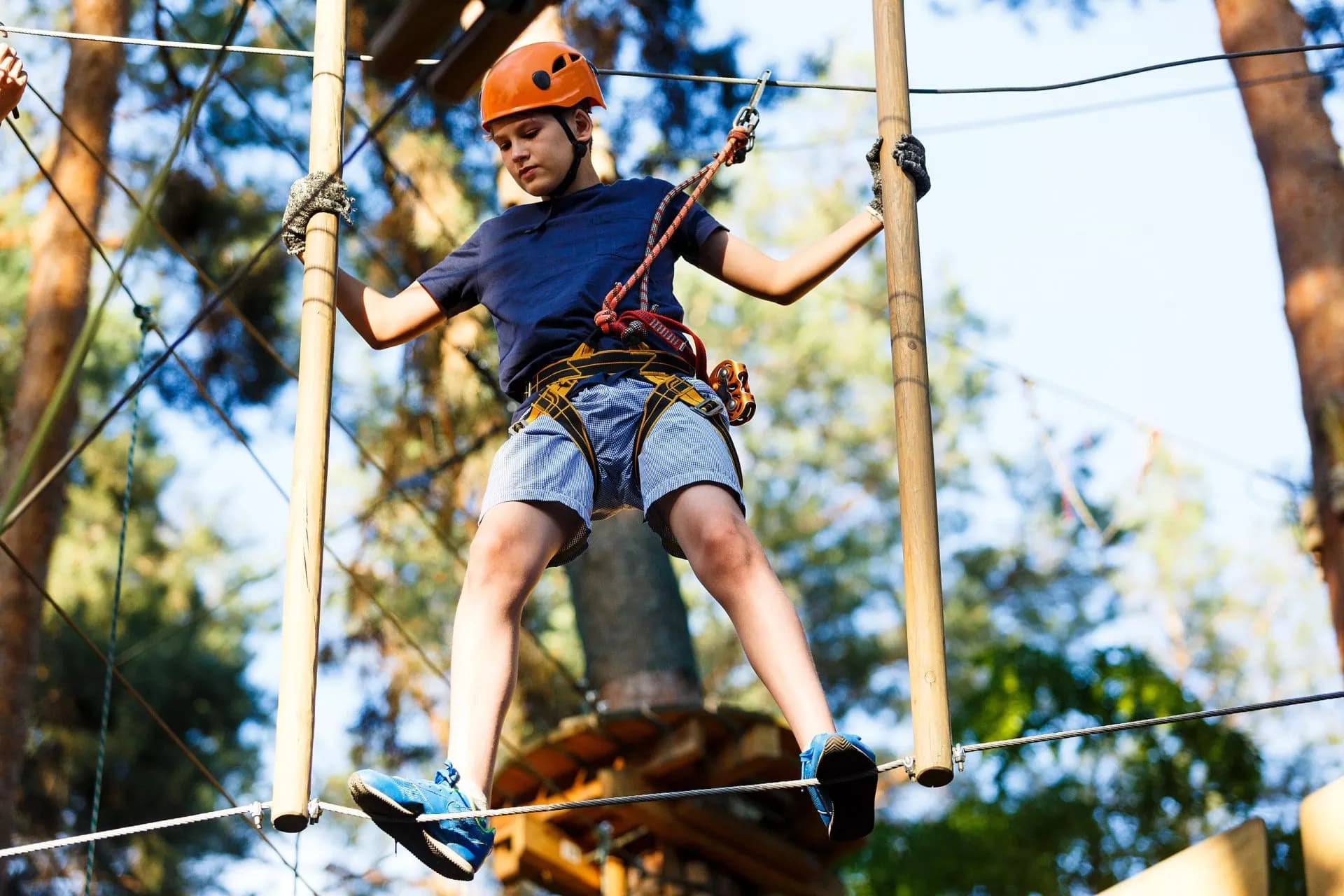 Boy in helmet traversing high ropes course obstacle in sunny forest adventure park.