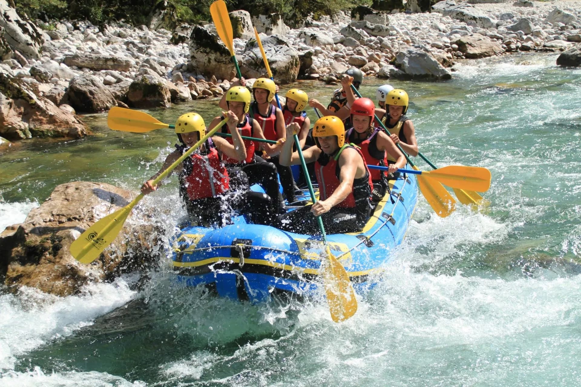 White water rafting group paddling blue raft through rapids by rocky river bank in Soca River.