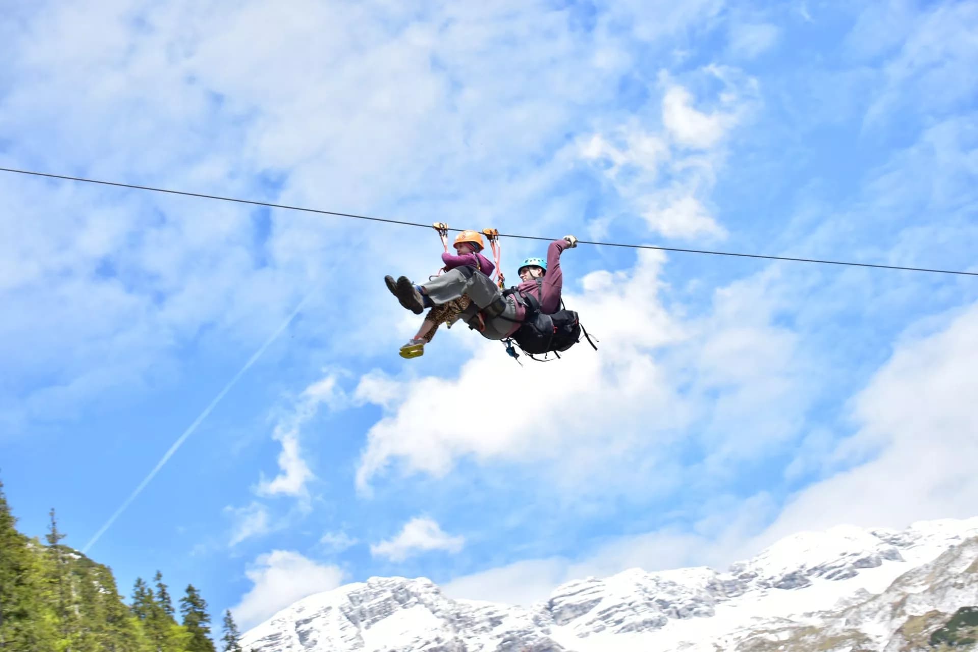 Tandem zip-lining over snow-capped mountains and green slopes under a blue, cloudy sky.