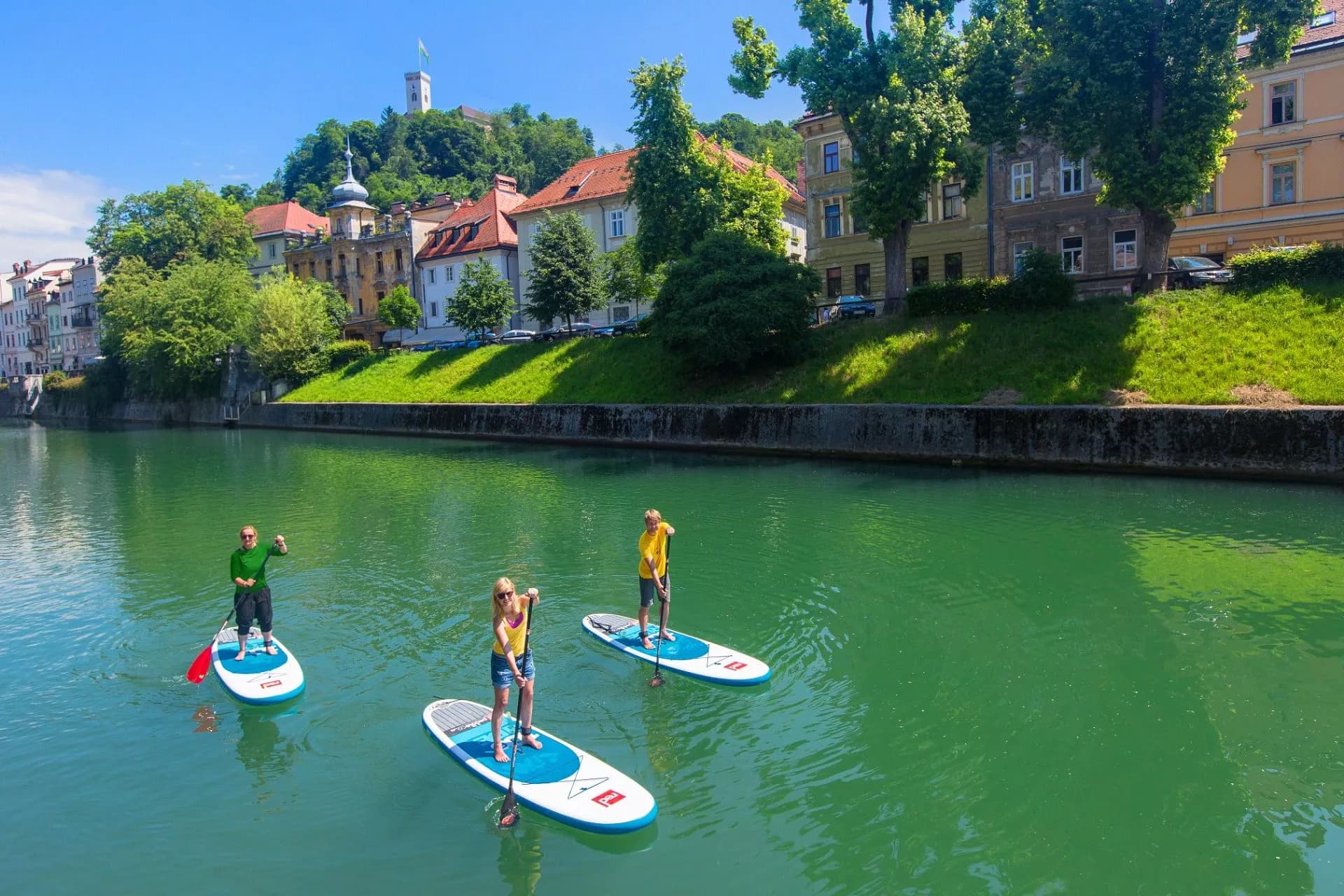 Stand-up paddleboarding on green river near Ljubljana Castle and historic buildings.