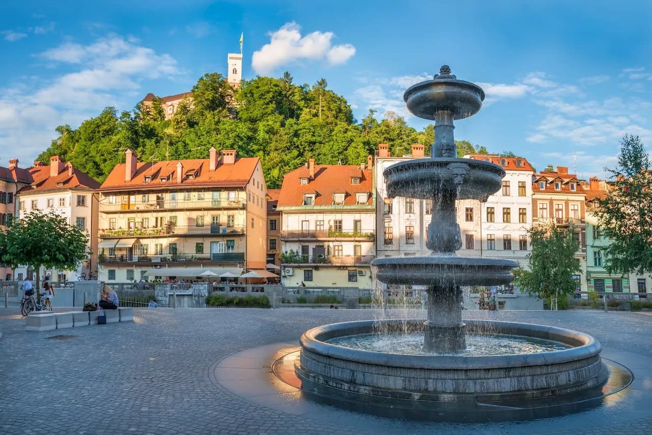 Fountain in Ljubljana City Center square with Ljubljana Castle on wooded hill.