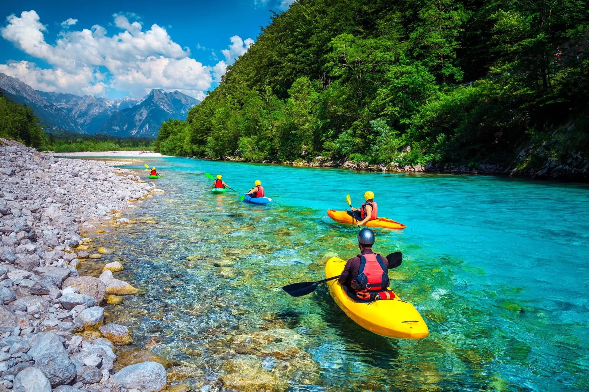 People kayaking on the clear turquoise Soca River with rocky banks and forested mountains.