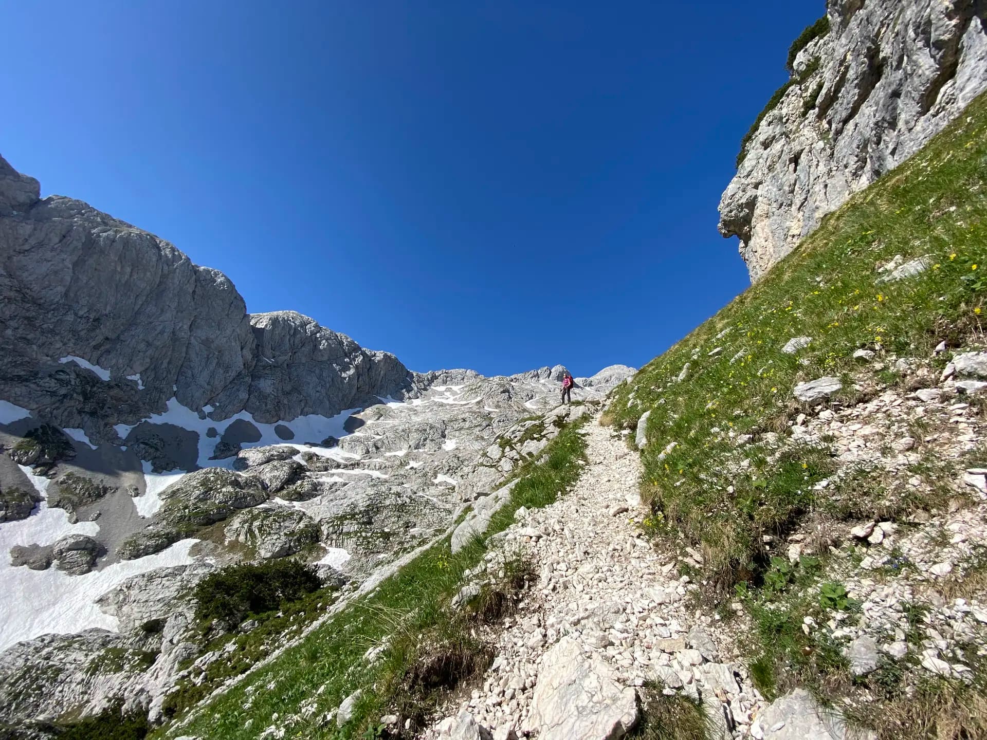 Hiker on a rocky mountain trail surrounded by steep limestone peaks and patches of snow.