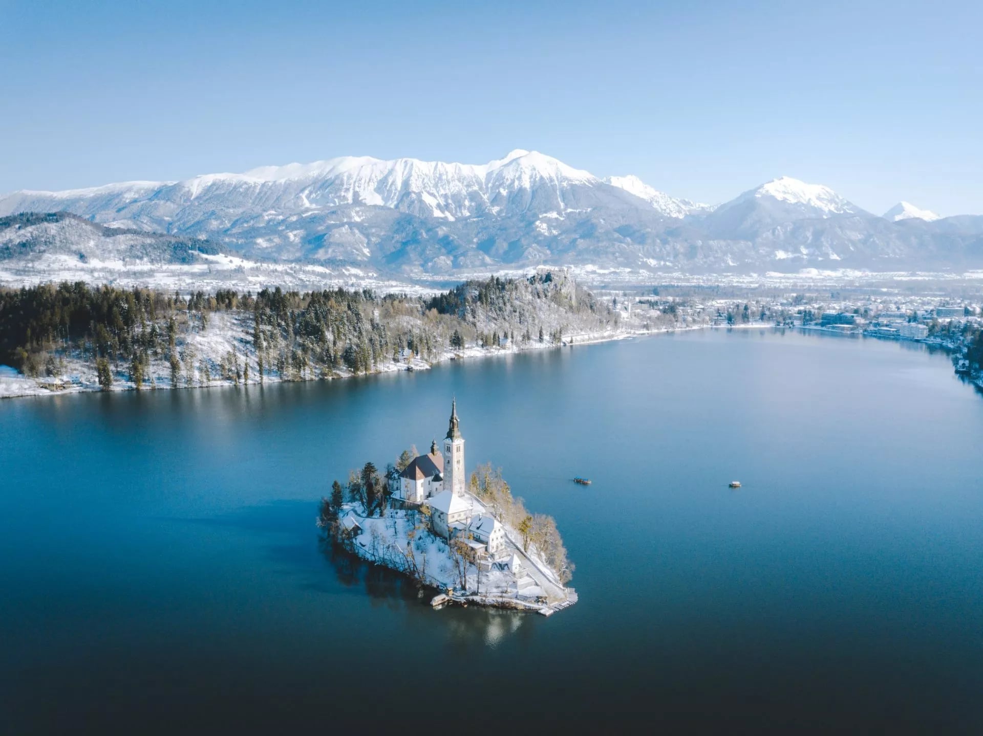 Aerial view of Bled Island church in winter on lake with snow-capped mountains in background.