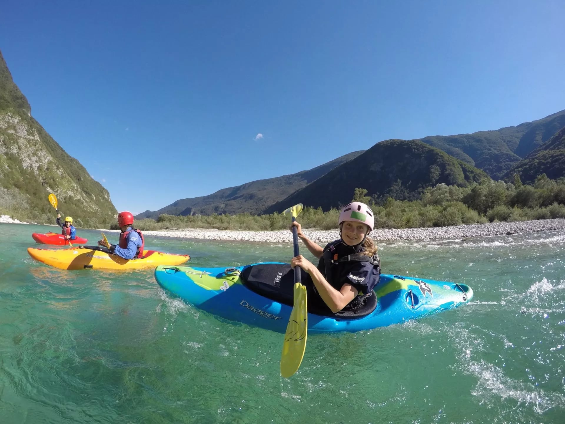 Family kayaking on turquoise river water with green mountains under a clear blue sky in Slovenia.