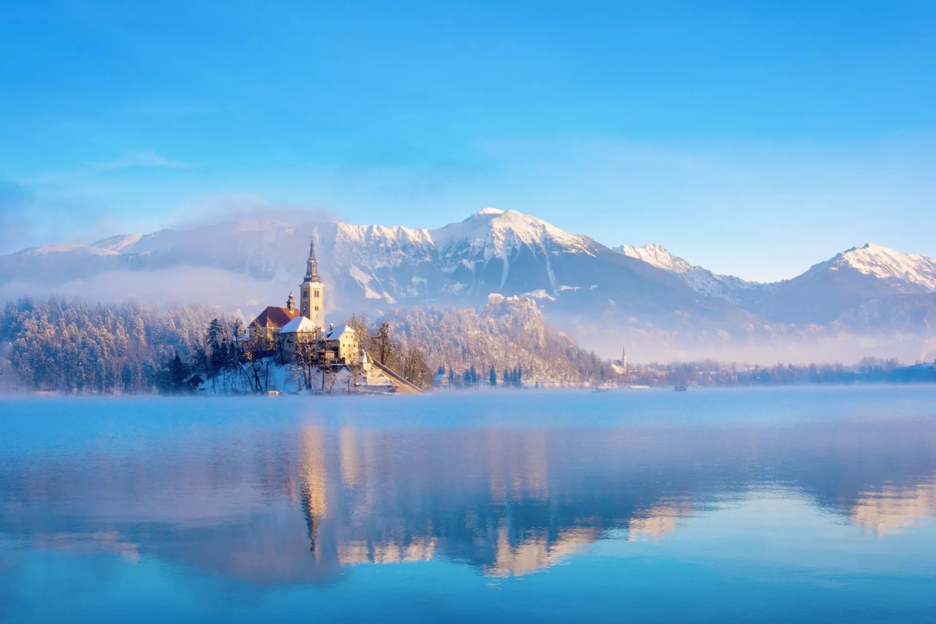 Lake Bled island church in winter with snow-capped mountains reflecting in the misty lake