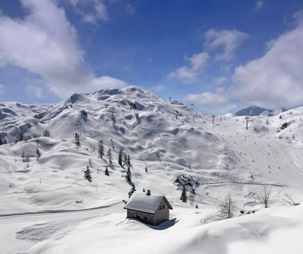 Small wooden cabin in deep snow at a ski resort with snow-covered mountains under blue sky.