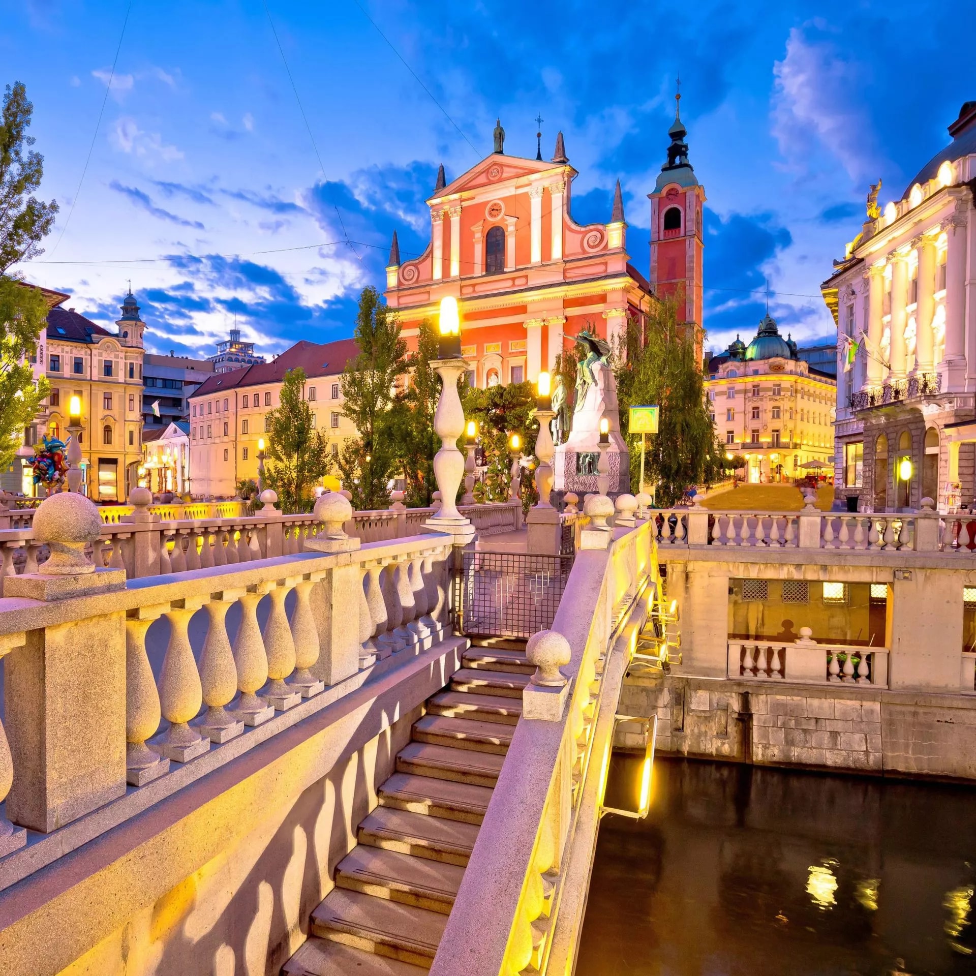 Ljubljana stone bridge steps leading to illuminated pink Franciscan Church at dusk.