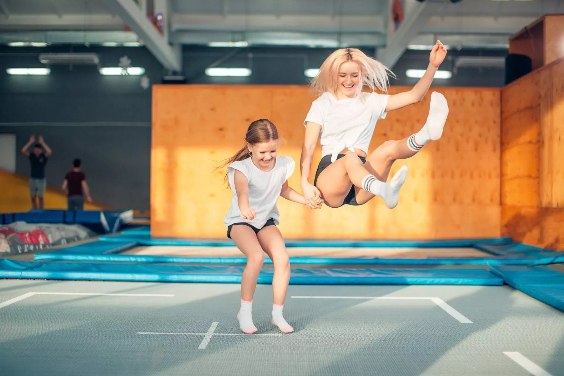 Two people jumping high while holding hands on trampolines inside a large indoor park.