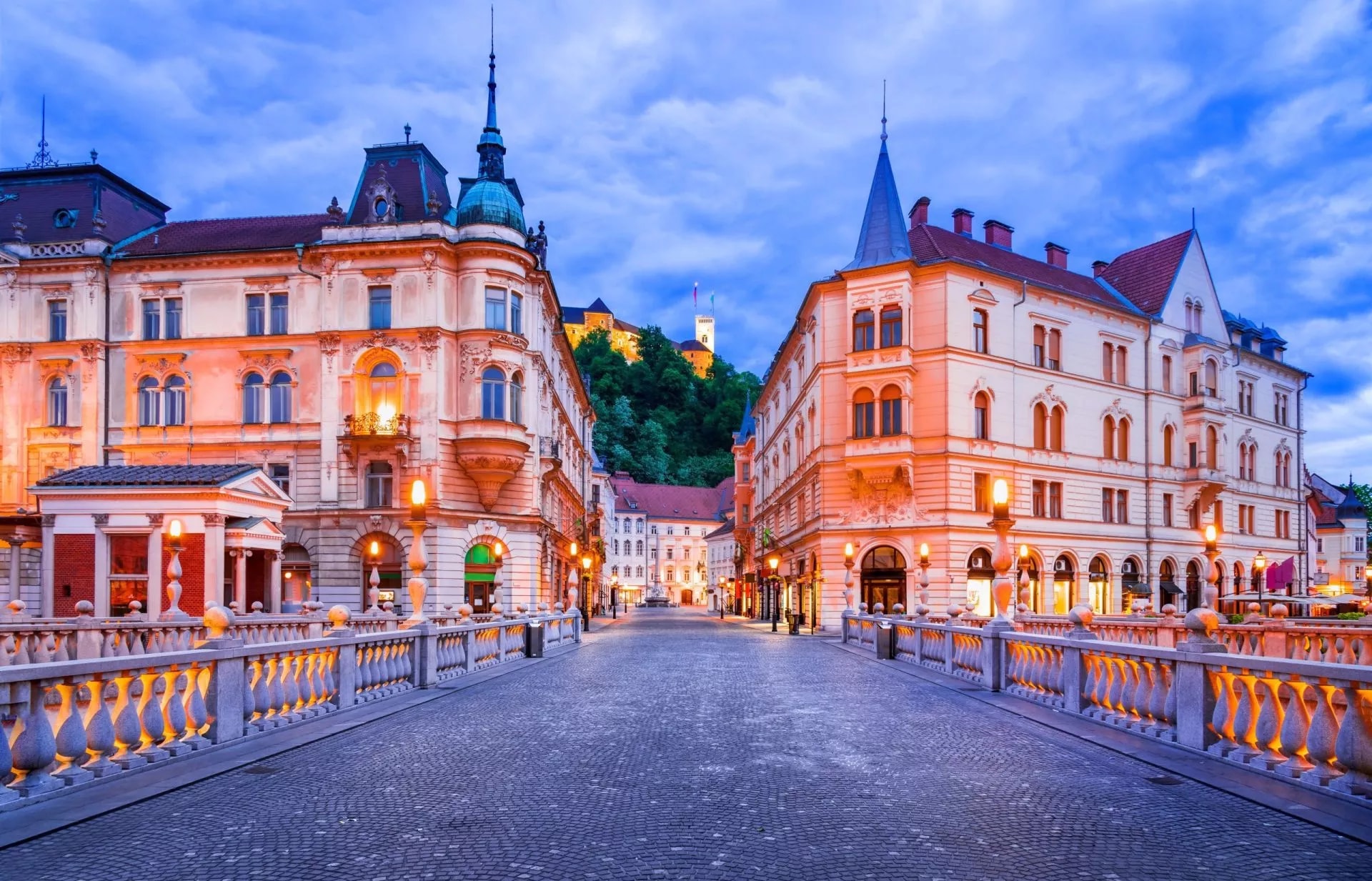 Cobblestone bridge leading toward Ljubljana Castle illuminated at dusk with historic buildings.