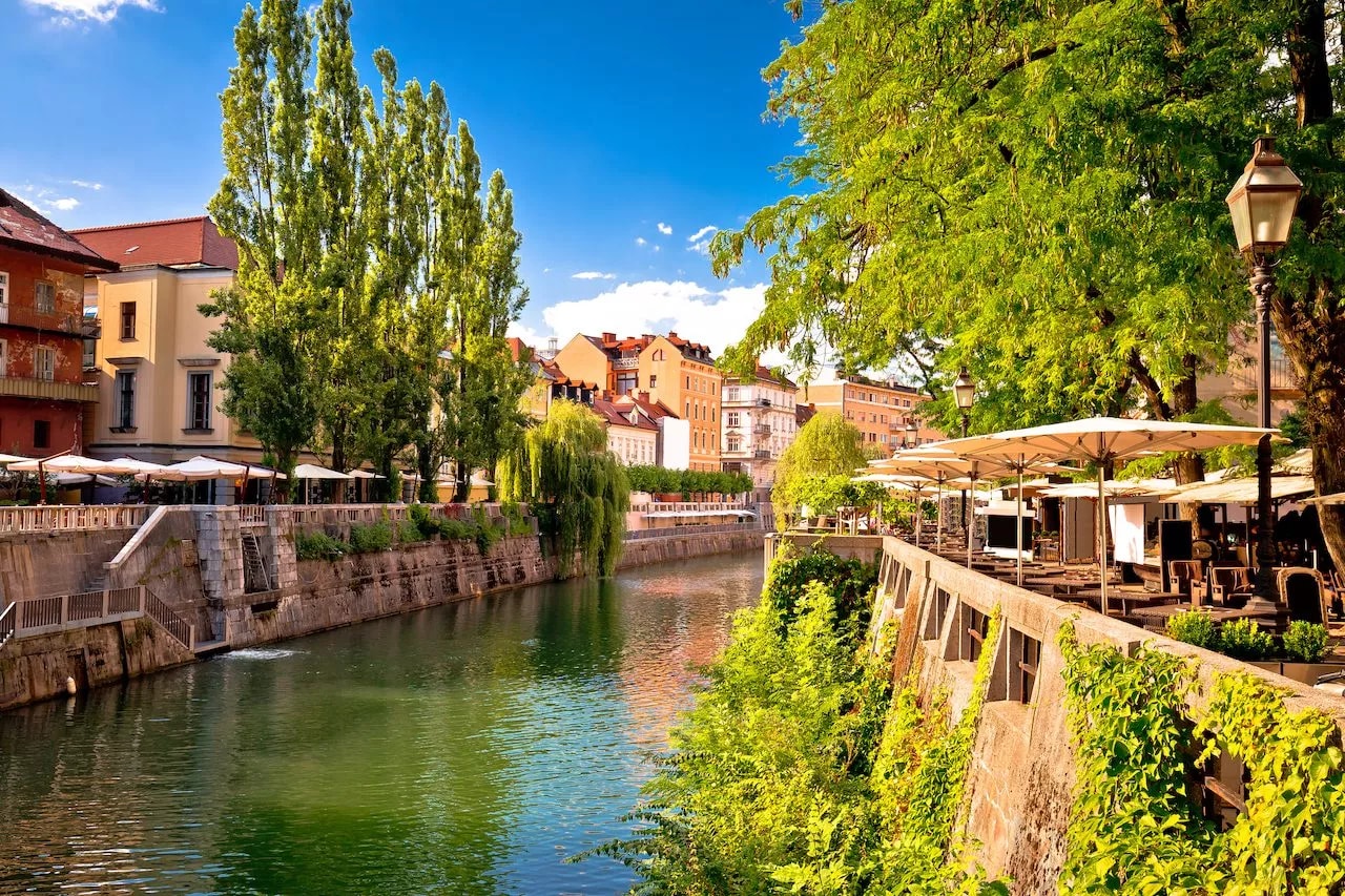 Riverfront outdoor cafe seating with umbrellas along a canal in sunny Ljubljana.
