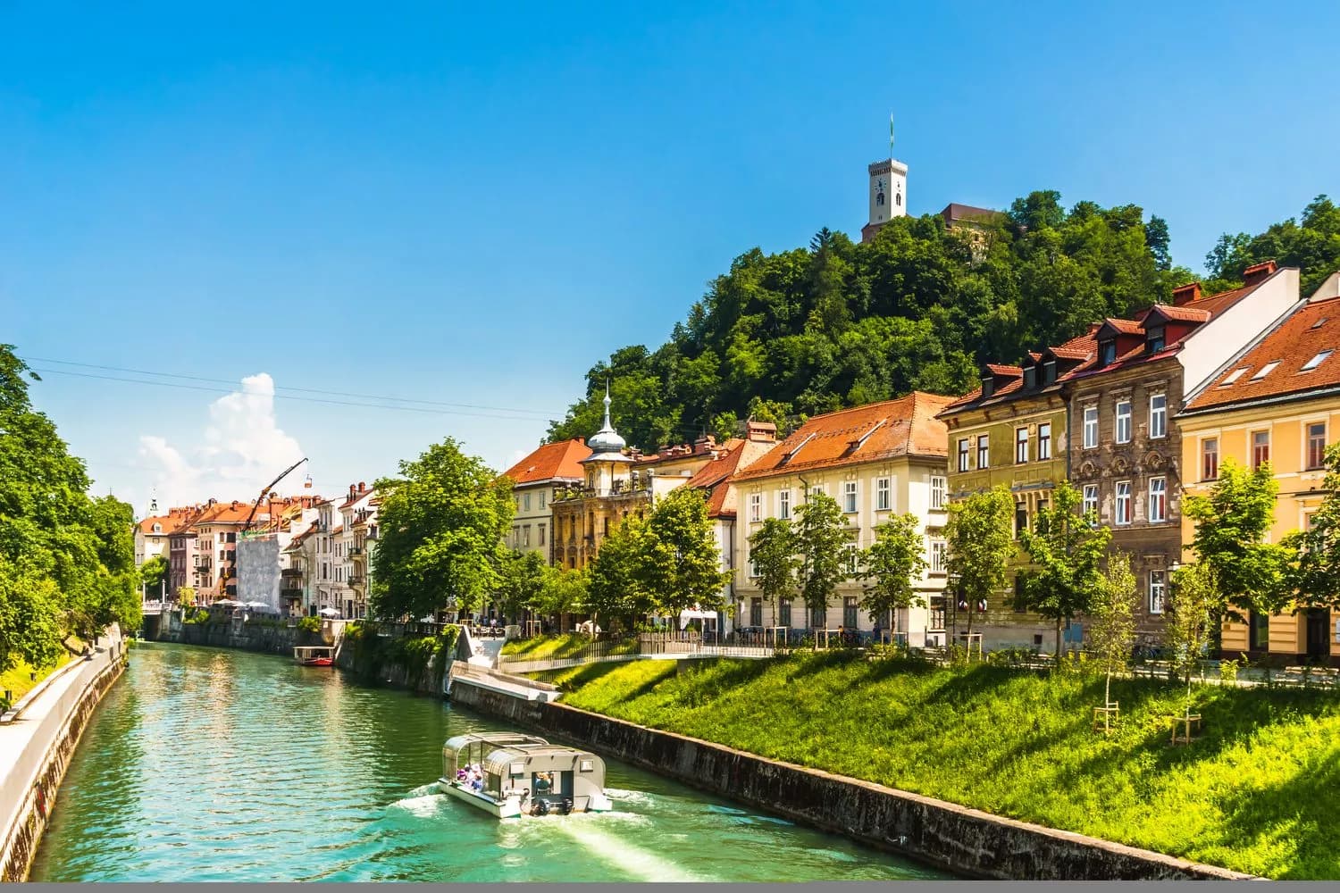 Tour boat on Ljubljanica River below Ljubljana Castle on a sunny day.