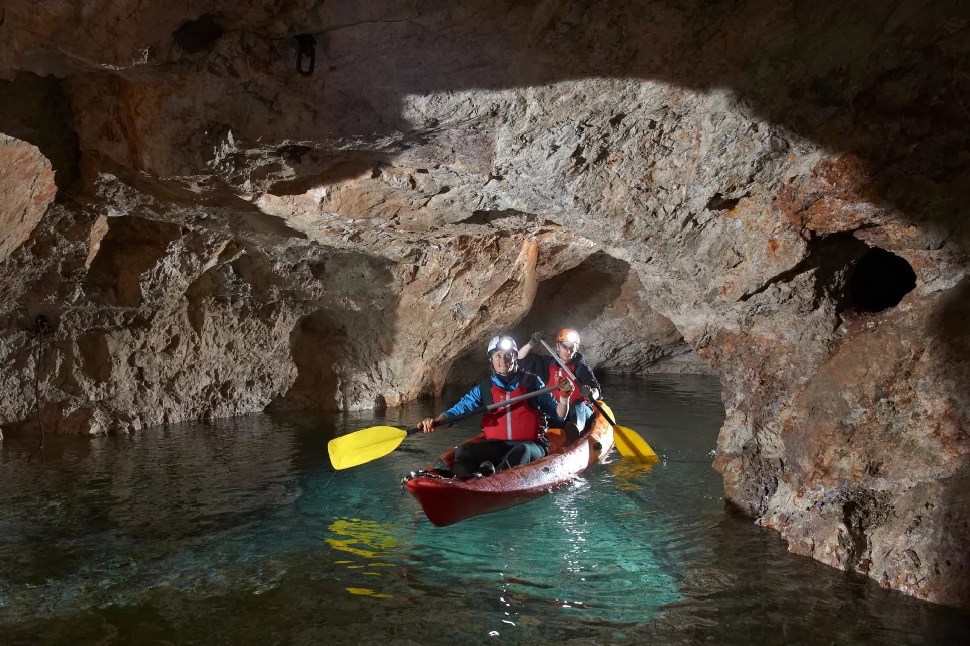 Kayaking with two people inside a red boat through a dark cave with clear water.