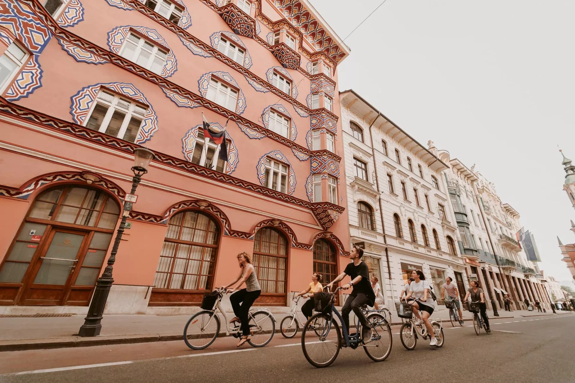 Cyclists riding past a pink building with ornate trim in Ljubljana city center.