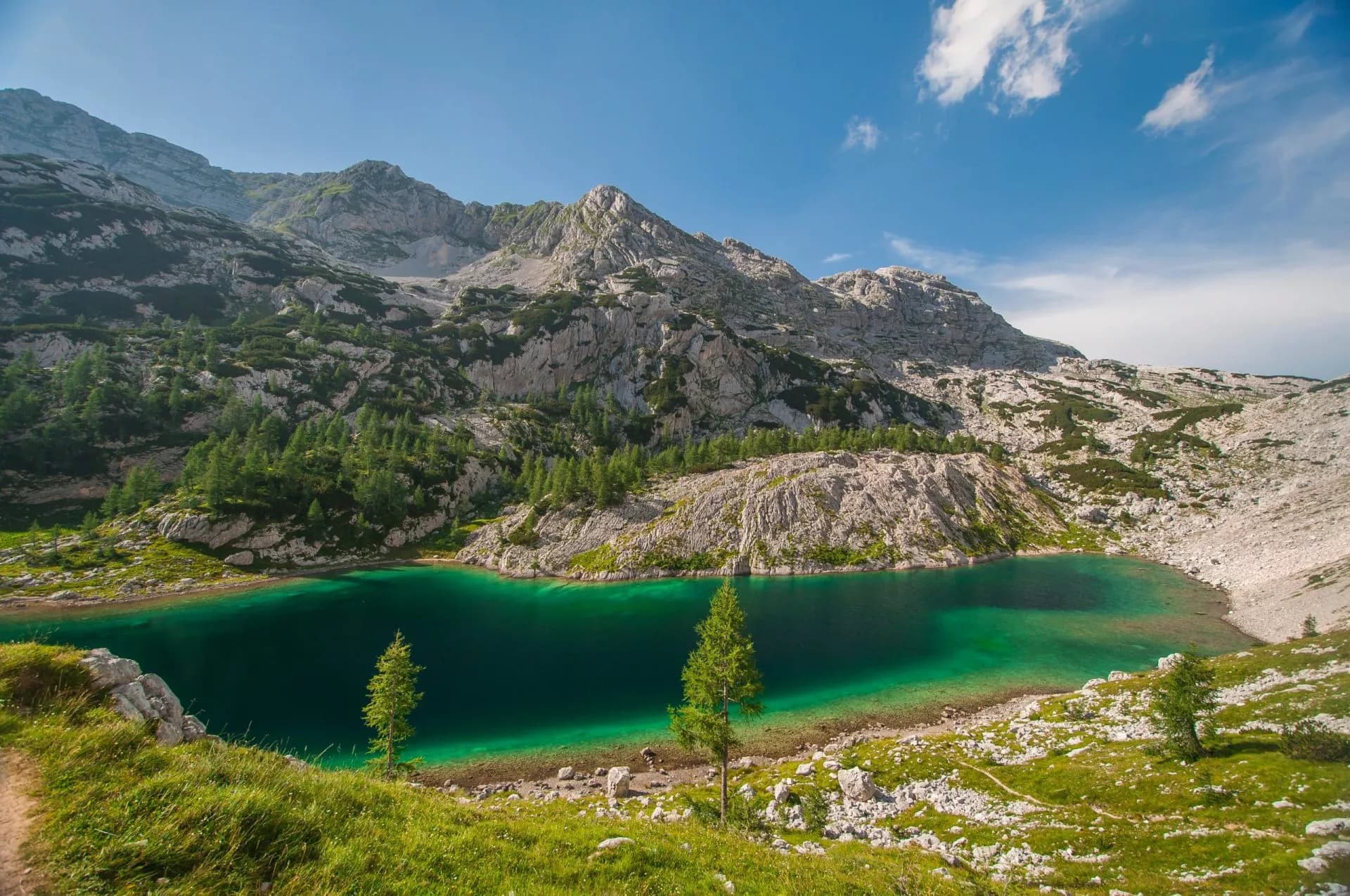 Alpine lake with emerald water surrounded by rocky mountains and green vegetation under a blue sky.