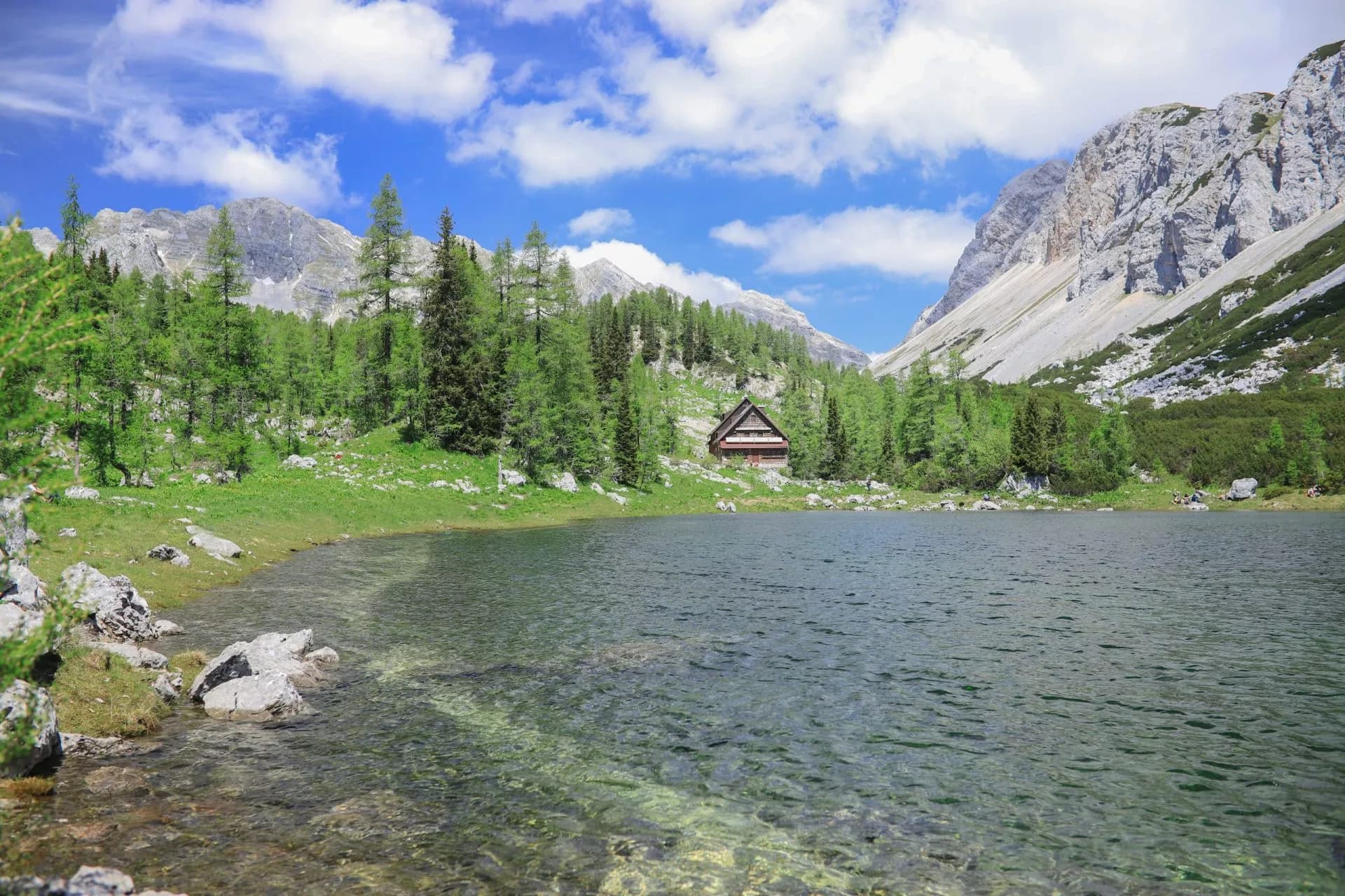 Hut at Triglav Lakes with clear water, green forest, and rocky mountains under blue sky.