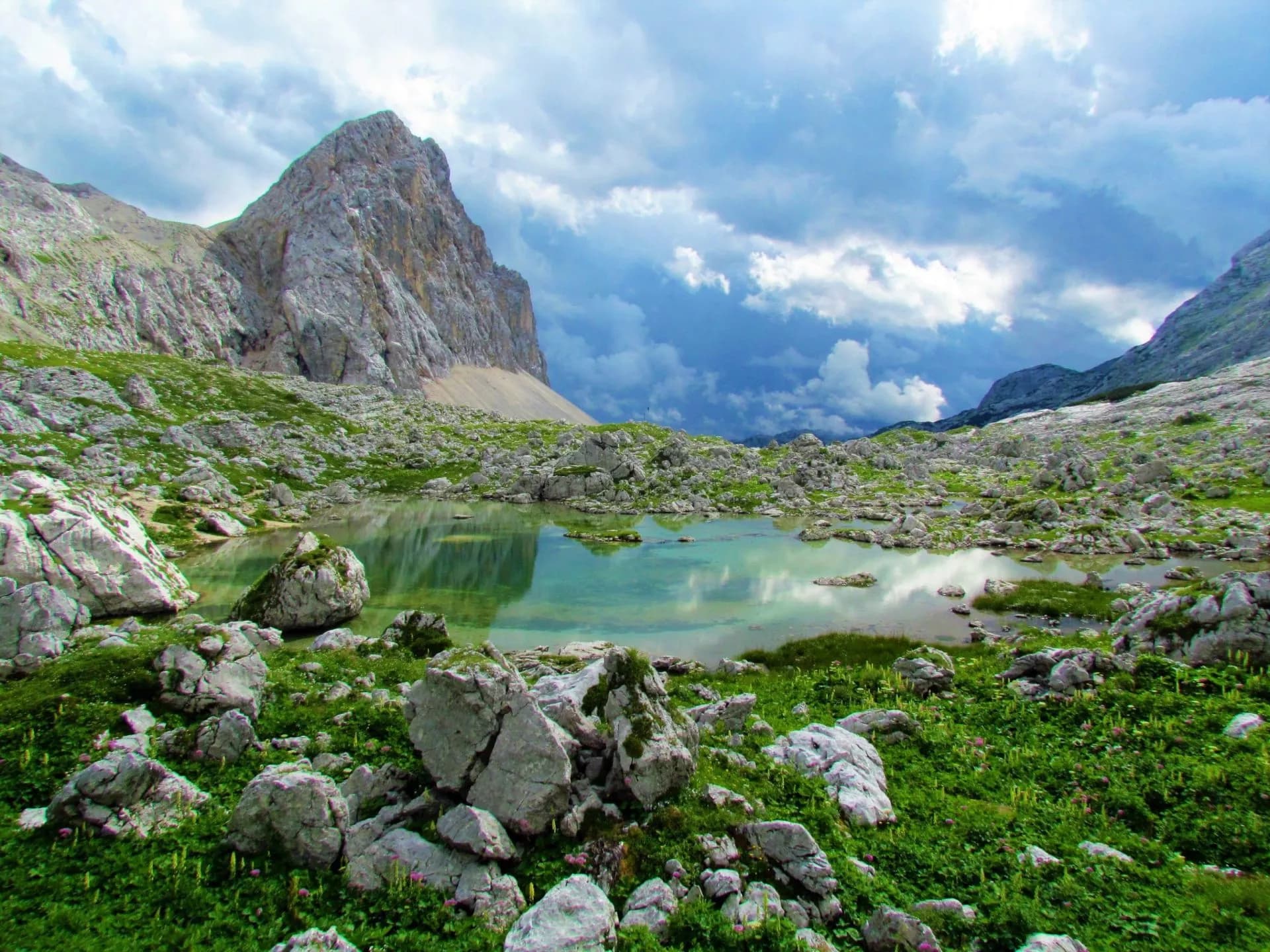 Alpine lake reflecting dramatic clouds, surrounded by rocky terrain and green summer foliage, Triglav.