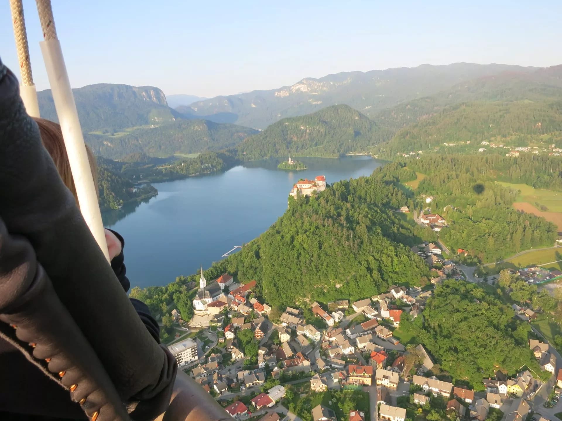 Hot air balloon view of Lake Bled, castle on cliff, and island church surrounded by mountains.