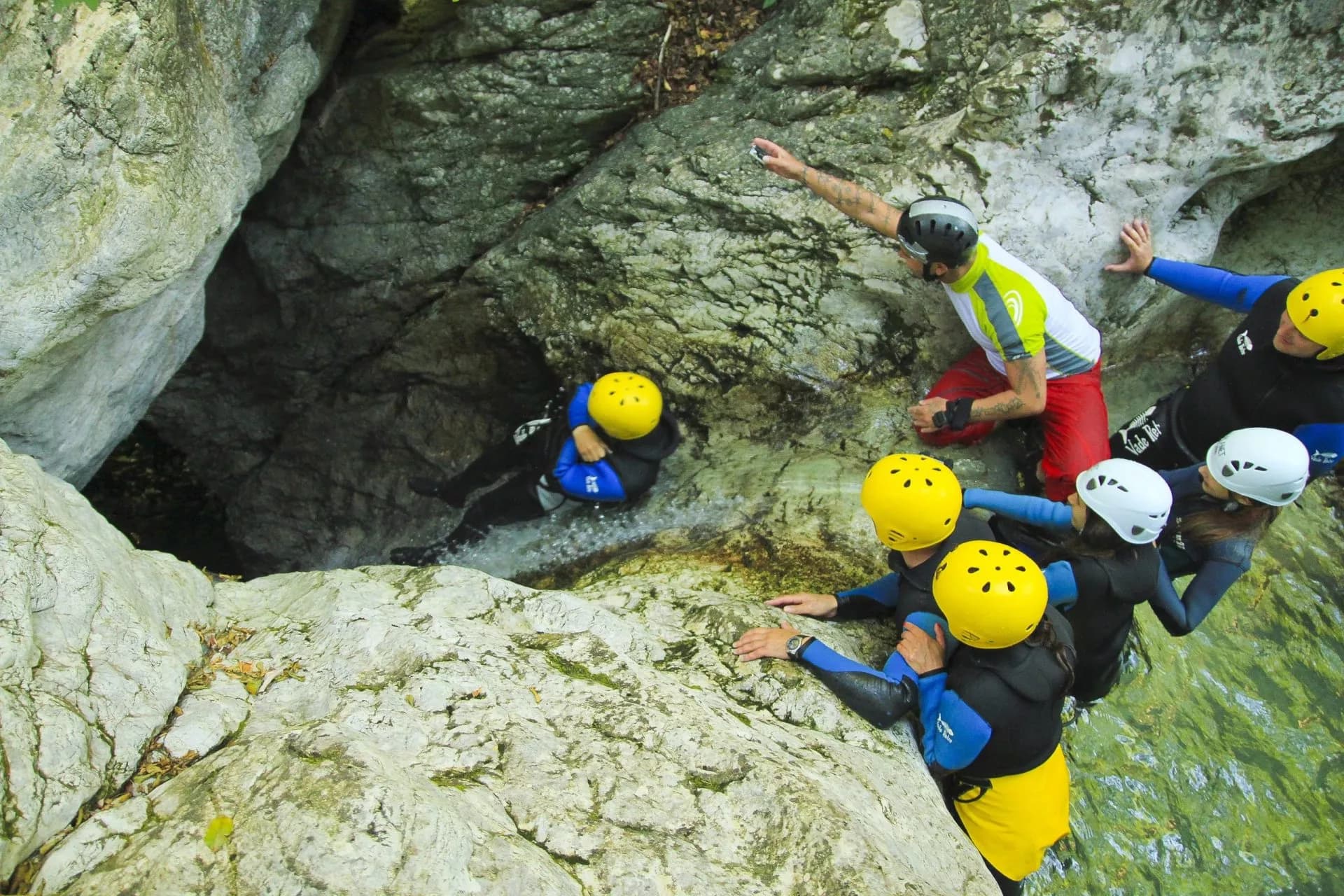 Canyoning group with helmets descending steep, wet rocks into a narrow gorge with rushing water.
