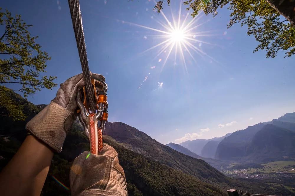 Ziplining with gloved hands gripping the cable high above forested mountains under a bright sun.