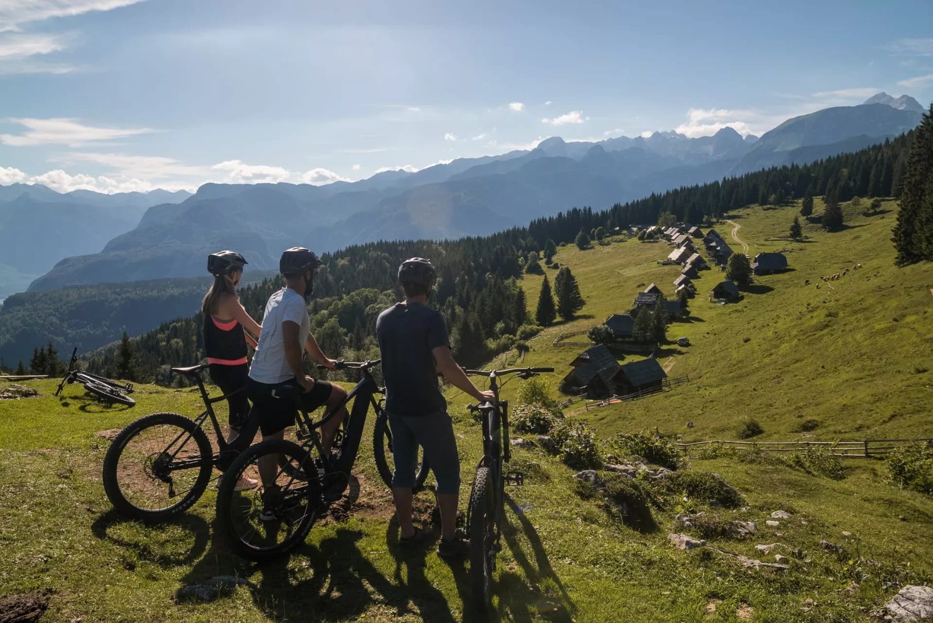 Cyclists with mountain bikes overlooking a green alpine meadow with wooden huts in Zajamniki, Pokljuka.