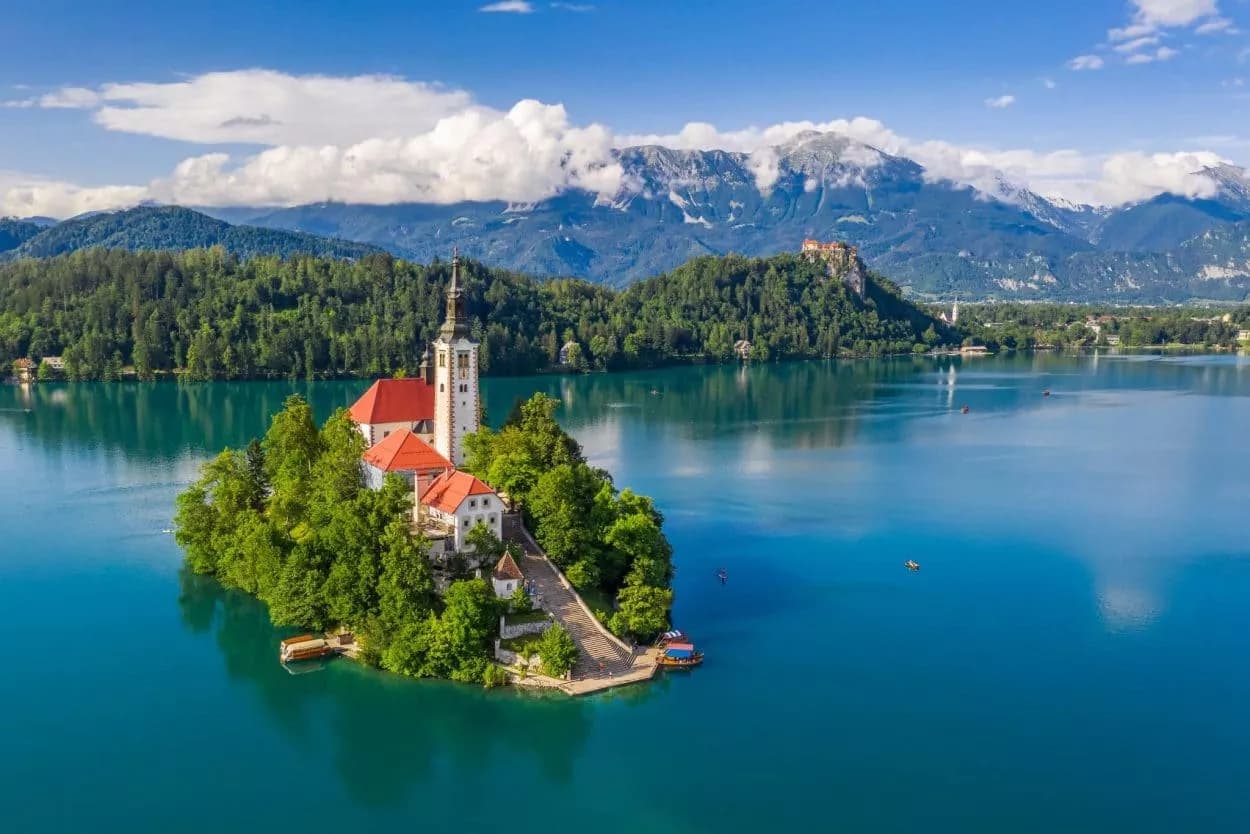 Church on island in Lake Bled with mountains and castle on shore under blue sky