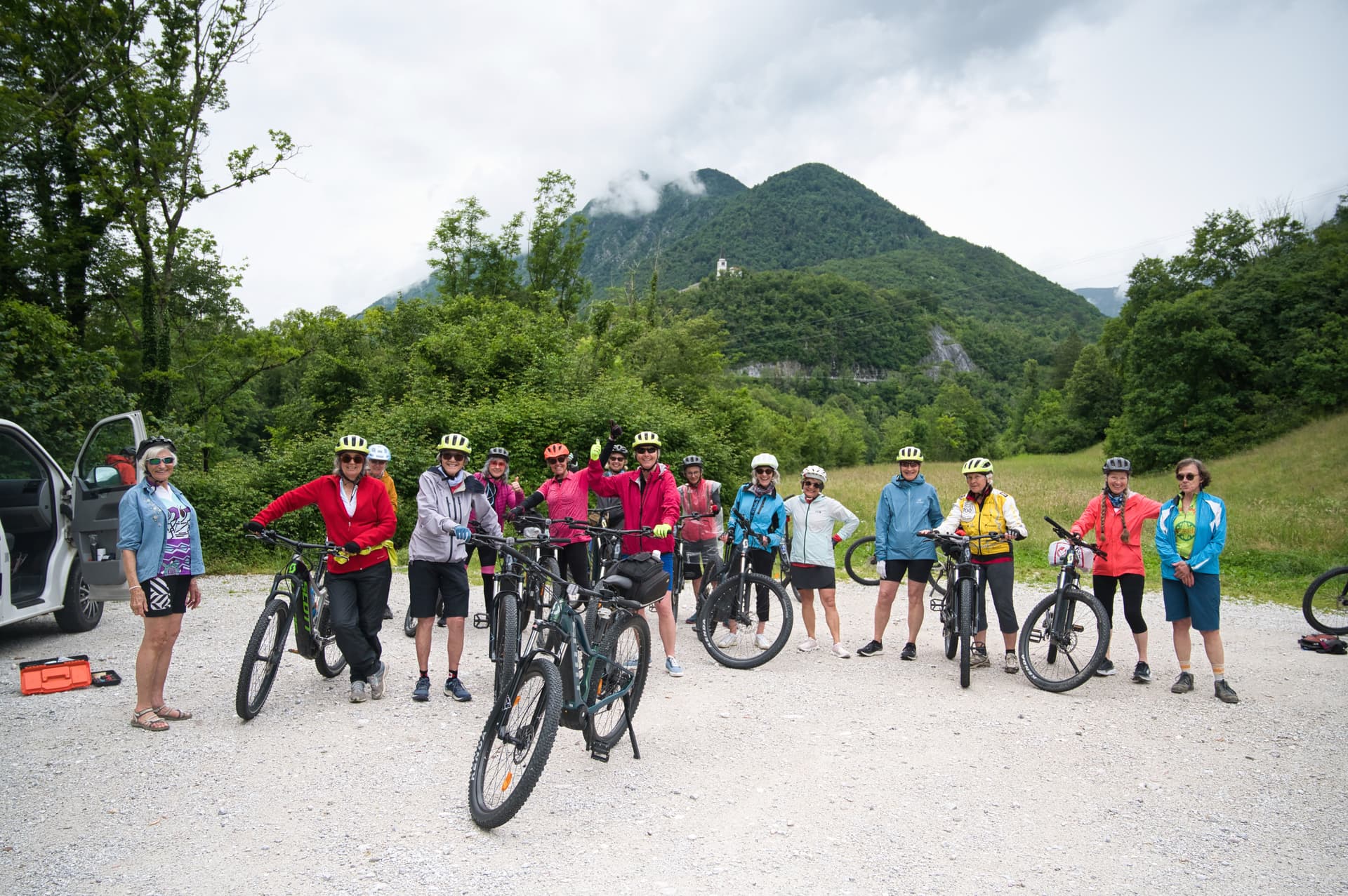 Cyclists posing with mountain bikes on gravel road near Julian Alps and forested hills.