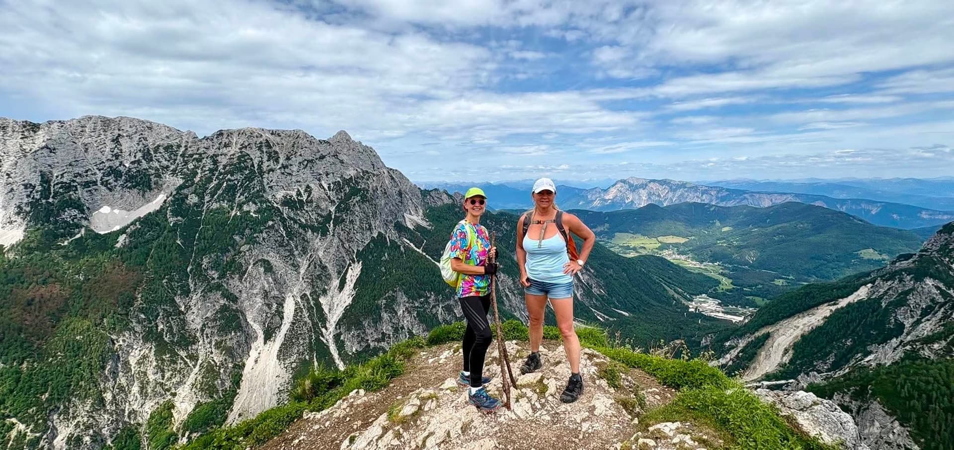 Two hikers standing on a mountain summit ridge with panoramic alpine views